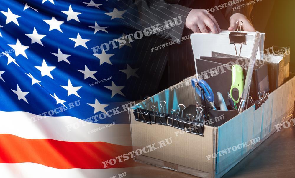 Фотография показывает коробку с офисными принадлежностями и документами, находящуюся в руках человека на фоне флага США. Это изображение символизирует увольнение и проблему безработицы в стране.